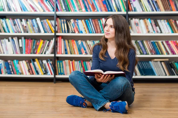 Улыбающаяся молодая девушка сидит на полу в библиотеке с — стоковое фото