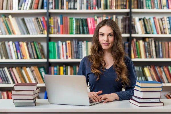 Sorridente ragazza seduta ad una scrivania in biblioteca a lavorare con — Foto Stock