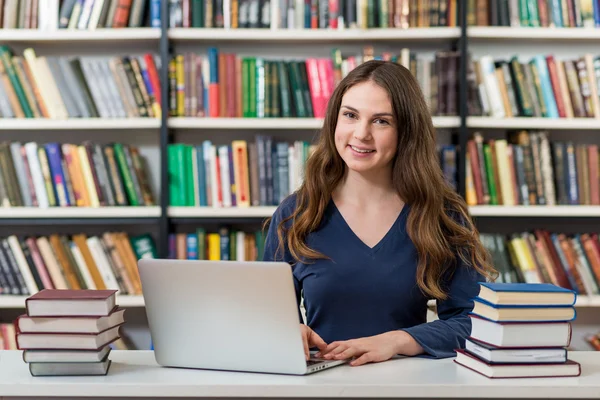 Усміхнена молода дівчина сидить за столом у бібліотеці, з якою працює — стокове фото