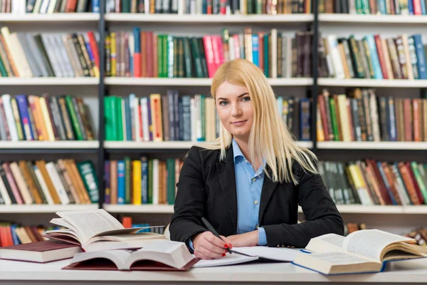 Lächelndes junges Mädchen mit blonden Haaren sitzt an einem Schreibtisch in der Bibliothek — Stockfoto