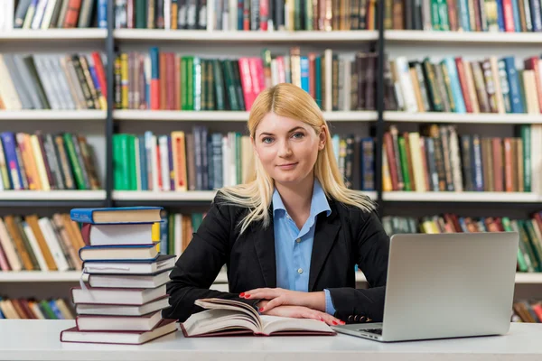 Усміхнена молода дівчина сидить за столом у бібліотеці, з якою працює — стокове фото