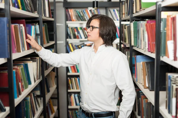 Молодий чоловік з темним волоссям вибирає книгу, що стоїть між стелажем — стокове фото