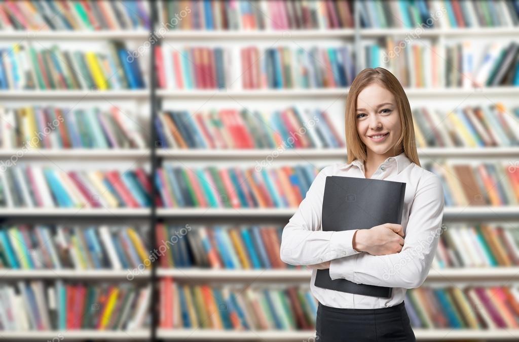 young woman standing, holding notebook in front of book shelves