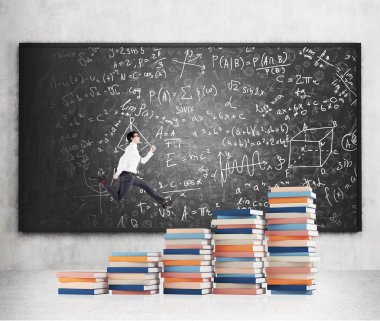 Young man with a folder in hand running up stairs made of piles of books of different size, blackboard with notes at the concrete background.