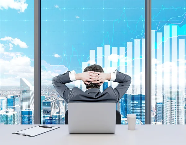 Rear view of a relaxing businessman who is sitting with his back to the laptop looking into the window with semi-transparent graphs on it. — Stockfoto