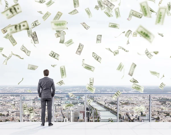Young businessman with hands in pockets standing on the roof of a business centre and looking at New York. Money falling from above. — Stock Photo, Image