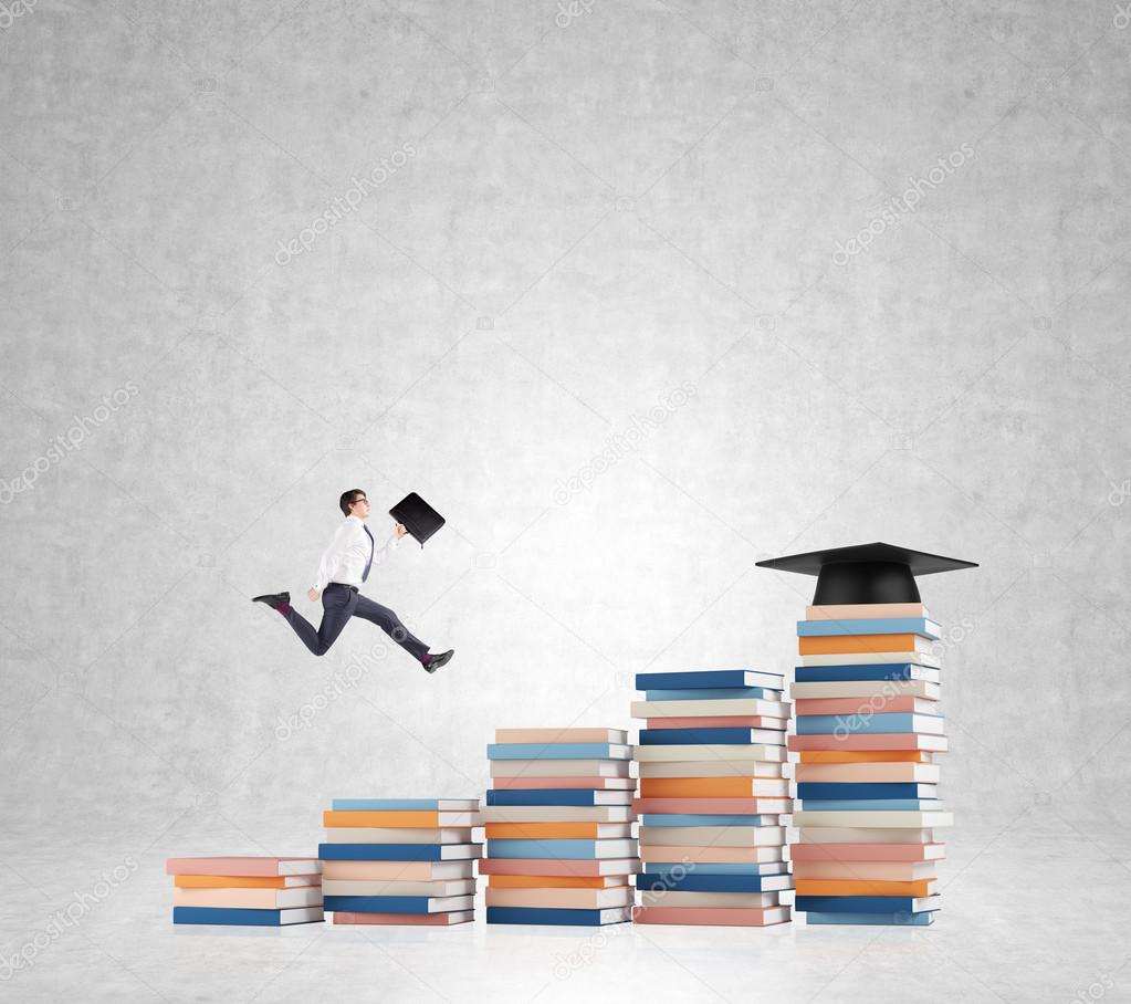 Young man with a folder in hand running up stairs made of piles of books of different size, graduation hat on the highest, concrete background.