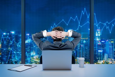 Rear view of a relaxing trader who is sitting in front of a window, laptop behind him.
