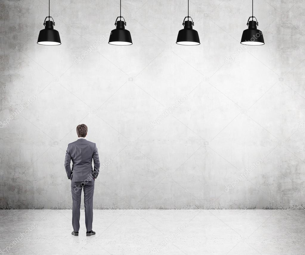 Young businessman standing with his hands in pockets front of a concrete wall, five lamps above.