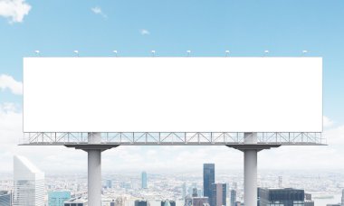 A huge blank billboard on two supports, city view and blue sky at the background.