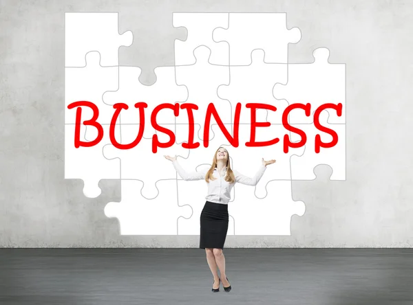 A young businesswoman looking up and smiling with hands raised standing in front of a puzzle with a word 'business' on a concrete wall. — Stockfoto
