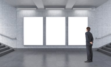 man with hands at back standing to the right of three blank vertical billboards in the unerground.
