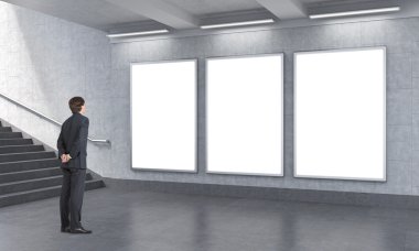 man with hands at back standing to the right of three blank vertical billboards in the unerground.