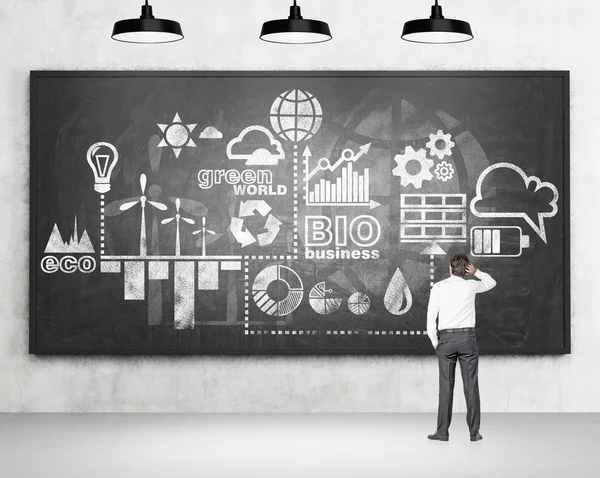 Man with a hand on head standing in front of a scheme of clean energy production drawn on a blackboard. — Stock Fotó