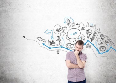 A young man with hand to the chin thinking and standing in front of a concrete wall with many different business icons drawn on it over a graph.