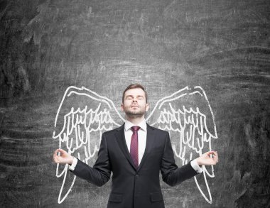 A young man standing in a posture of meditation in front of a blackboard with angel wings painted on it as if they were his.