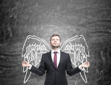 A young man standing in a posture of meditation in front of a blackboard with angel wings painted on it as if they were his. clipart