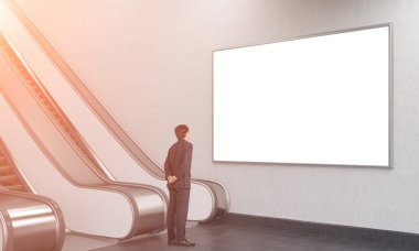 young businessman with his hands at the back looking at a blank poster over an escalator in the underground.