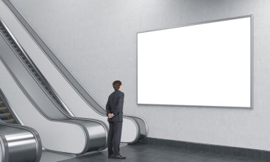 young businessman with his hands at the back looking at a blank poster over an escalator in the underground.