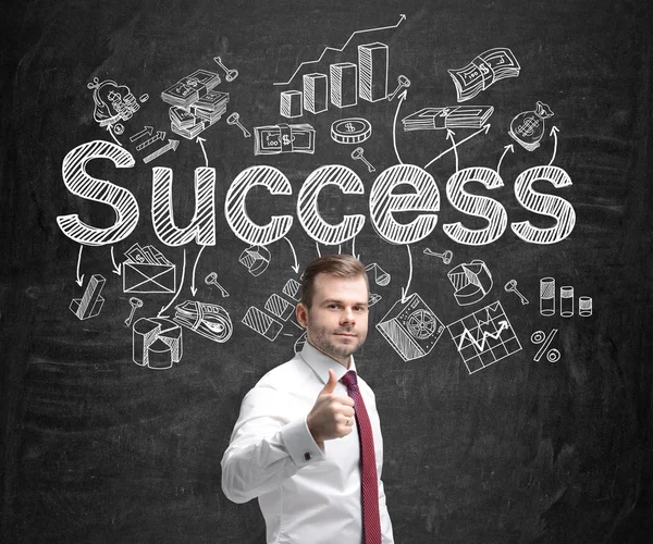 A young man holding a thumb up and standing in front of a blackboard with many different business icons and the word 'success' drawn on it. Concept of successful business development. — 图库照片