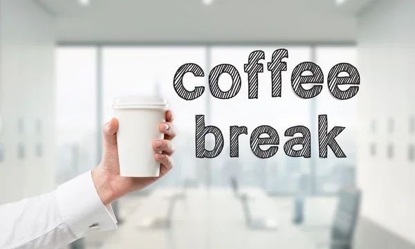 Una mano sosteniendo una taza de papel. "Pausa para el café" escrito a la derecha. Oficina borrosa en el fondo . — Foto de Stock