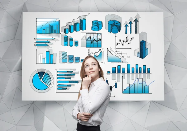 A young pretty businesswoman with hand to the chin looking up is standing in front of a white poster with many different blue graphs drawn on it. — Stockfoto