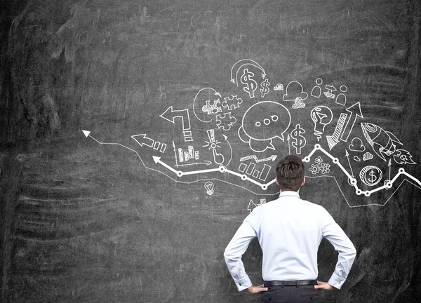 A young man with hands on hips standing in front of a blackboard with different business icons over a graph drawn on it. — Stock fotografie