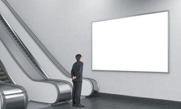 Jovem empresário com as mãos na parte de trás olhando para um cartaz em branco sobre uma escada rolante no subterrâneo . — Fotografia de Stock
