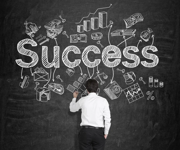 A young man drawing many different business icons and the word 'success' on the blackboard. — Stockfoto