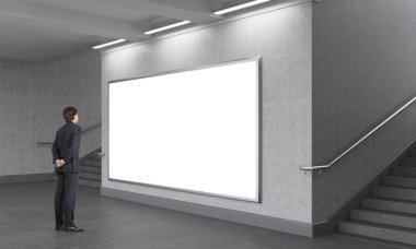 A man standing in front of a big blank horizontal billboard in the underground, stairs up on both sides.