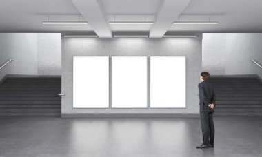 A man standing in front of three blank vertical billboards in the underground, stairs up on both sides.