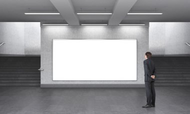 A man standing in front of a big blank horizontal billboard in the underground, stairs up on both sides.