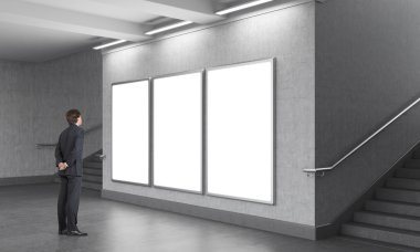 A man standing in front of three blank vertical billboards in the underground, stairs up on both sides.