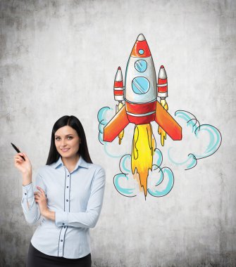 A young woman with a pen in hand standing in front of a concrete wall with a coloured image of a rocket to the right.