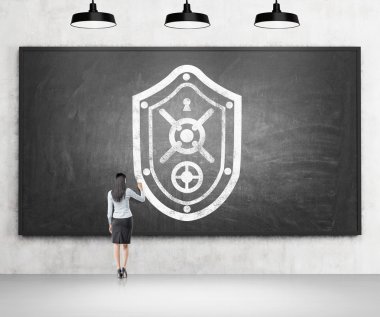 A woman drawing a shield resembling a safe door on the blackboard, four black lamps above.