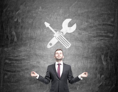 A businessman standing in the posture of meditation, a screwdriver and a wrench crossed are drawn on a blackboard over his head.