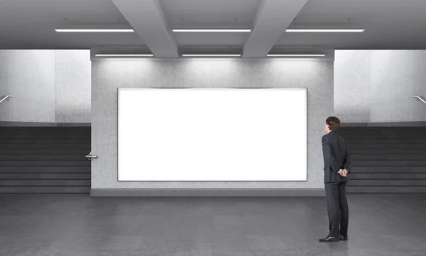 A man standing in front of a big blank horizontal billboard in the underground, stairs up on both sides. — Stockfoto