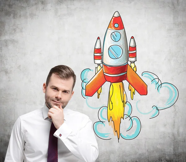 A young man with a hand to the chin standing in front of a concrete wall with a coloured image of a rocket to the right