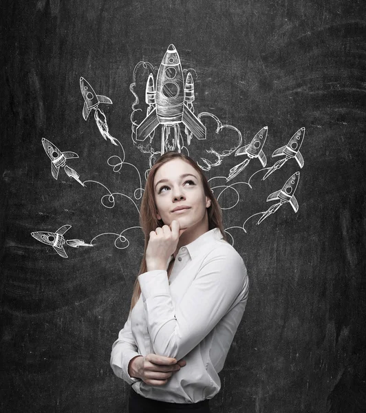 A smiling young woman standing at a black wall and looking up and aside, painted rockets flying from over her head. — Stockfoto