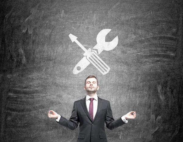 Ein Geschäftsmann steht in der Haltung der Meditation, ein Schraubenzieher und ein gekreuzter Schraubenschlüssel sind auf eine Tafel über seinem Kopf gezeichnet.. — Stockfoto