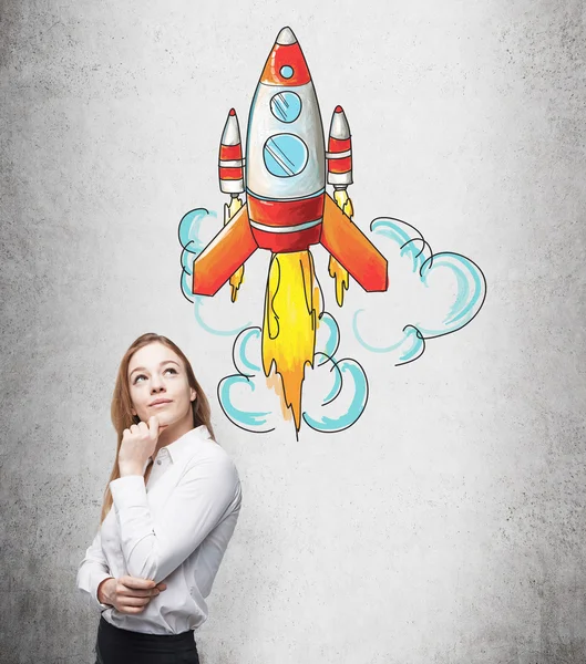 A smiling young woman standing at a concrete wall and looking up and aside, a coloured painted rocket flying from over her head. — Stockfoto