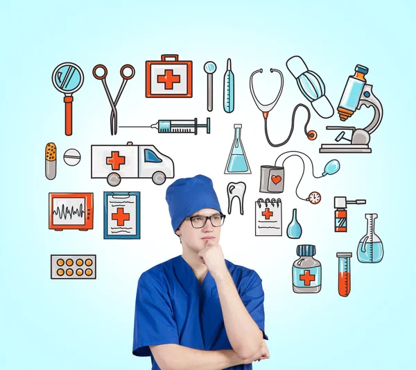 A young male doctor in a blue uniform with a hand on his chin standing in front of the blue wall, many medical icons drawn on it. — Stockfoto