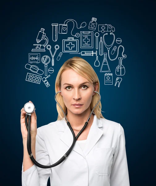 A young female doctor in a white smock holding a phonendoscope and standing in front of the dark blue wall, many medical icons drawn on it. Front view. — Stockfoto