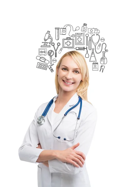 Una joven doctora con una bata blanca con las manos cruzadas de pie frente a la pared blanca, muchos iconos médicos dibujados sobre su cabeza . —  Fotos de Stock