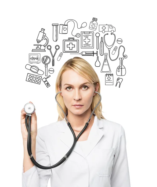 A young female doctor in a white smock holding a phonendoscope and standing in front of the white wall, many medical icons drawn on it. — Stockfoto