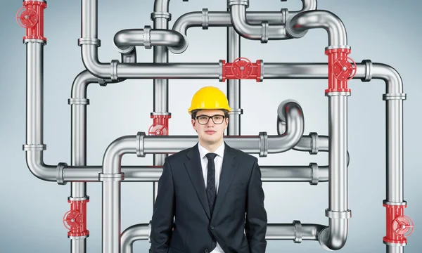 A businessman in a suit and a yellow helmet looking in front of him, a pipeline with red taps behind him. Light blue background. — Stockfoto