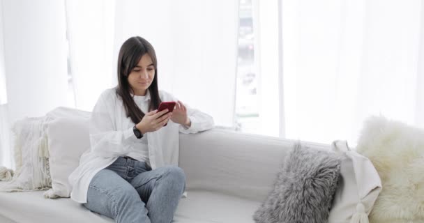 若い女性は家でソファに座っているスマートフォンのメッセンジャーでチャットしています. — ストック動画