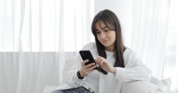 Happy young woman chatting in messenger on smartphone sitting on sofa at home. — Stock Video