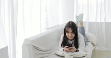 Genç bayan akıllı telefonuyla haberciyle sohbet ediyor. Evdeki kanepede yatıyor..