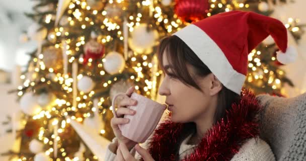 Retrato de mujer feliz en sombrero rojo está bebiendo té cerca del árbol de Navidad en casa. — Vídeo de stock